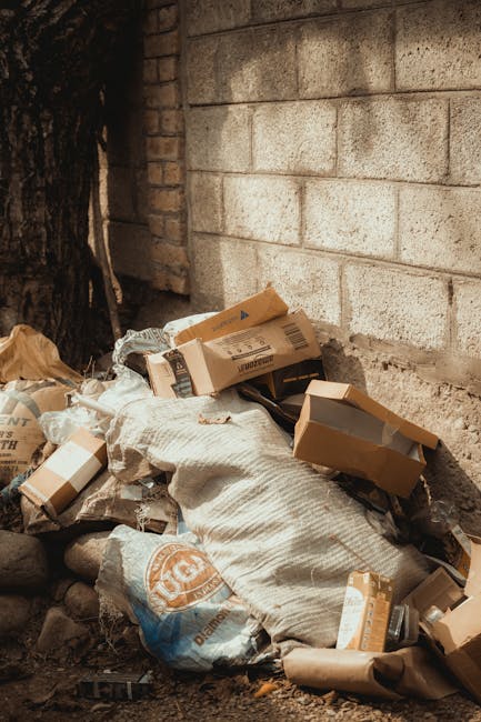 A pile of discarded cardboard boxes, paper bags, and packaging materials is gathered at the base of a rough brick wall and concrete block wall, partially sheltered by a large tree trunk on the left side of the image. The packaging includes flattened and partially crumpled boxes in various shades of brown and grey, some with printed labels or barcodes visible. Among the debris is a large, cream-colored sack or bag, possibly filled with waste or rubble, resting on a bed of small rocks and dirt. The scene appears to be outdoors, with natural sunlight illuminating the area, highlighting the textures of the cardboard and the surface of the wall behind. This image exemplifies a typical instance of excess packaging waste that might be handled through private rubbish removal services such as those offered by Rubbish Removal Roehampton, reflecting on the importance of proper waste disposal and alternative collection methods for items that are not suitable for curbside rubbish collection.