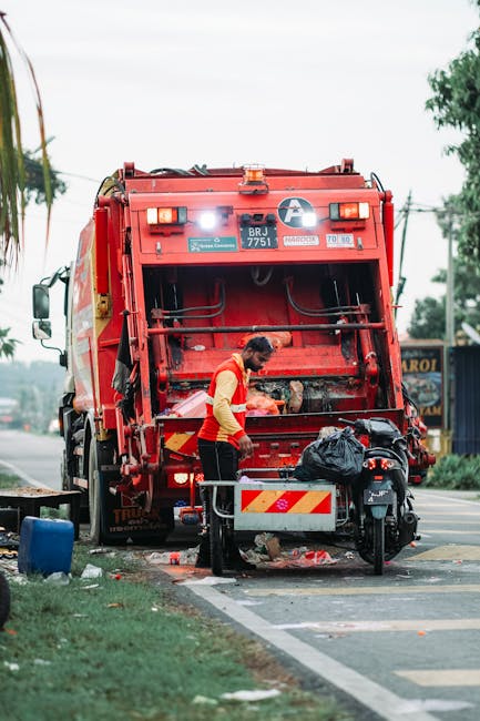 A municipal waste collection worker operates a large red rubbish collection truck on a street. The worker, dressed in a red and yellow high-visibility vest, stands next to the open rear loading area of the truck, which is equipped with hydraulic mechanisms and safety railings. The back of the truck is empty, suggesting recent collection activity, and a black plastic bag and some waste materials are visible near the worker. The scene takes place on a paved street with a sidewalk and a grassy verge, with trees and a few commercial buildings in the background. Overhead power lines are also visible, and the sky appears slightly overcast. The environment illustrates an instance of private waste collection, aligning with alternative rubbish removal methods, with the truck likely part of a local independent service such as Rubbish Removal Roehampton.