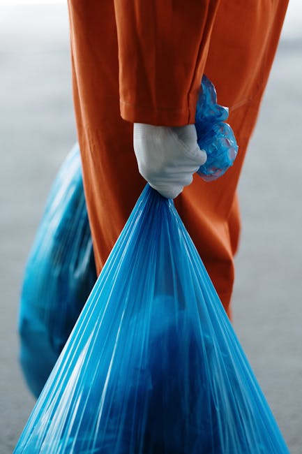 Close-up of a person's hand wearing a white glove, gripping a tightly tied blue plastic rubbish bag filled with waste, with a second similar bag visible in the background. The individual is dressed in an orange waterproof suit, indicating outdoor waste collection or disposal activities. The scene is set outdoors on a paved or concrete surface, with natural lighting suggesting daytime, and the focus is on the bags and hand, emphasizing the act of rubbish handling. This image relates to private waste management or rubbish removal services, illustrating the process of collecting and segregating waste materials for disposal or recycling, often associated with independent rubbish removal providers like Rubbish Removal Roehampton.